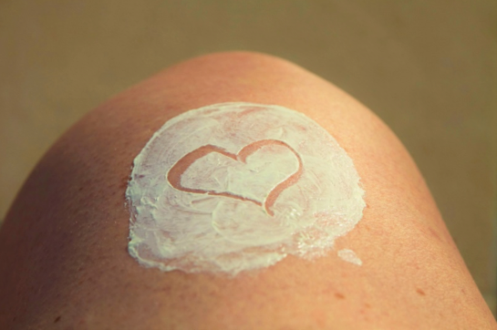 The image shows a close-up of a person's sun-kissed skin, likely a shoulder or knee, with a thick layer of white sunscreen applied in a circular patch - https://wmderma.com/