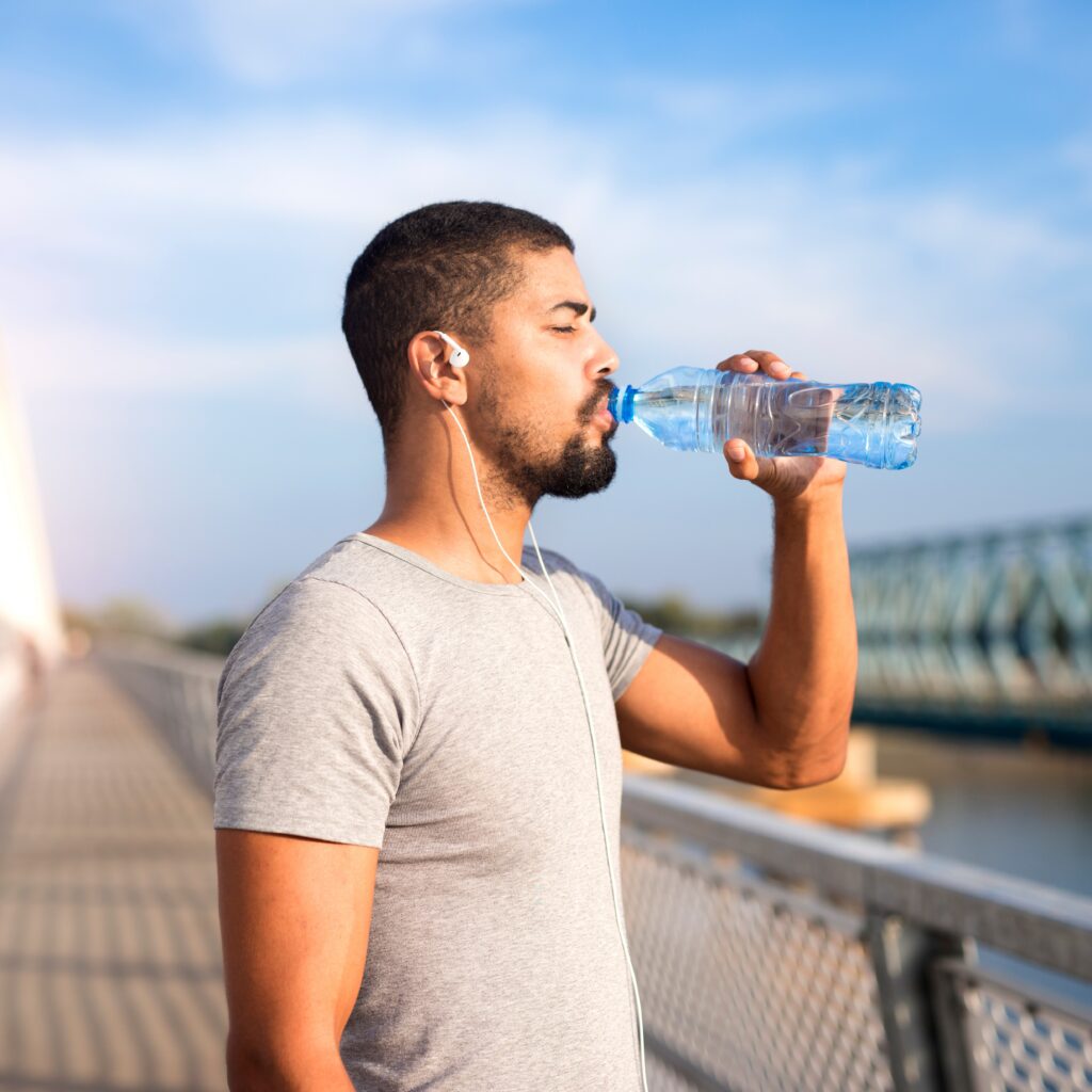 An athletic man with a beard and short dark hair is shown in profile, standing on a bridge and drinking from a clear plastic water bottle - https://wmderma.com/
