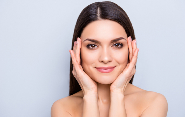 Smiling woman with clear skin framing her face with both hands, showcasing beauty and skincare results.