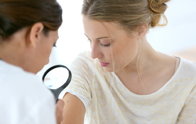 A healthcare professional examines a woman’s arm using a magnifying glass, possibly checking a skin condition or mole. 